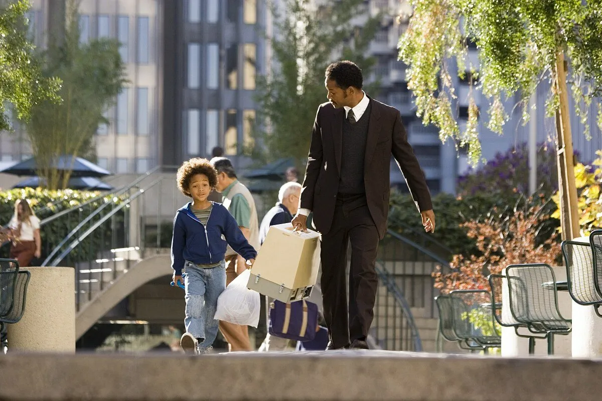 Post cover: Will Smith as Chris Gardner in The Pursuit of Happyness (2006) walks through a sunlit city plaza in a worn business suit, carrying a bulky medical scanner, while his young son walks beside him.